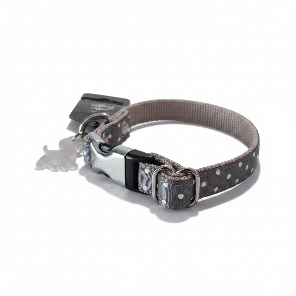 Un collier réglable pour petit chien, photographié de trois-quarts sur un fond blanc neutre. La sangle extérieure en tissu satiné est gris foncé avec des motifs à pois blancs, tandis que la doublure intérieure est en nylon tissé beige clair. Le collier est équipé d'une boucle à fermeture rapide en métal argenté et plastique noir, d'un passant de réglage métallique et d'un anneau en D auquel est fixé un petit pendentif transparent en forme de silhouette de chien.