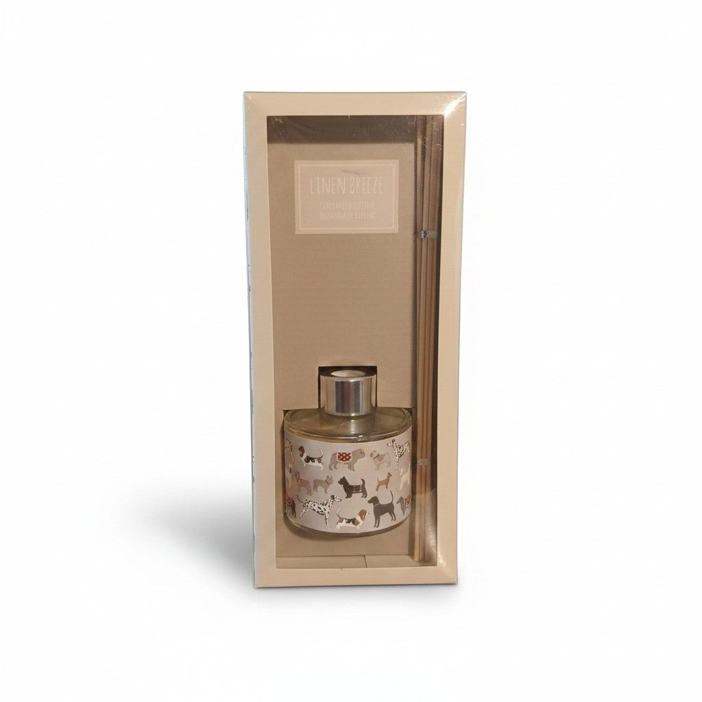 Un coffret rectangulaire beige contenant un kit de diffuseur de parfum d'ambiance, de face sur un fond blanc. Le coffret comprend un flacon en verre cylindrique rempli d'un liquide transparent, fermé par un bouchon argenté brillant. Le flacon est décoré  de nombreuses races de petits chiens (Dalmatien, Teckel, Carlin, Bouledogue). À droite du flacon, plusieurs longues tiges de rotin en bois naturel sont disposées à la verticale. Une étiquette  indique le parfum « LINEN BREEZE"