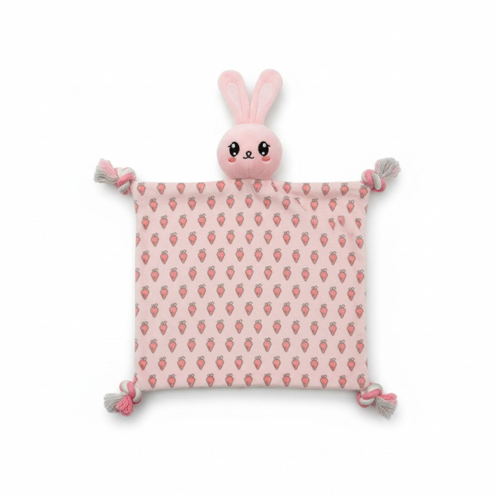 Doudou plat pour chien en forme de lapin rose pastel. Le corps en tissu présente de petits motifs imprimés de carottes oranges. La tête en peluche rembourrée surmonte le doudou, et les quatre coins se terminent par des nœuds en corde tressée blanc et rose.