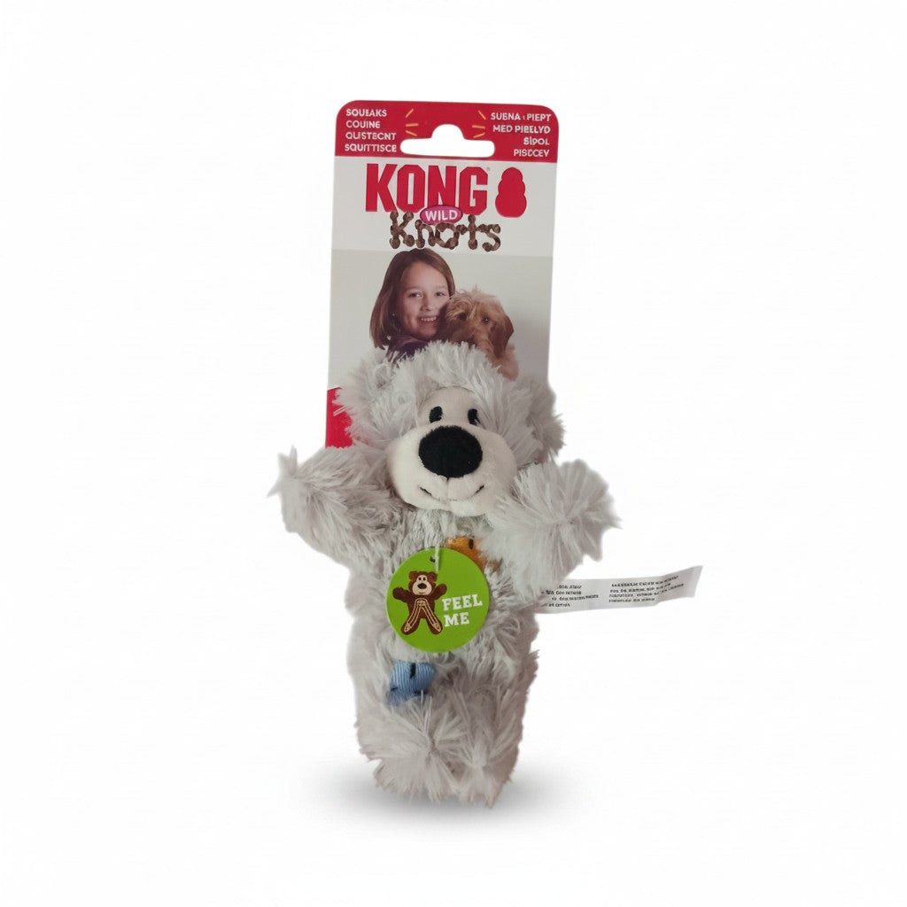 Peluche pour chien KONG Wild Knots Bear en version grise, présentée verticalement dans son emballage d'origine sur fond blanc. L'ours en fausse fourrure douce possède un museau blanc, un nez noir brodé et un corps souple. Une étiquette verte en forme d'ours avec l'inscription "Feel Me" est fixée sur sa poitrine. Le carton d'emballage supérieur est rouge et blanc.