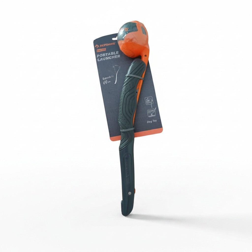 Lanceur de balle pliable pour chien de la marque SKIPDAWG, modèle "PORTABLE LAUNCHER", présenté plié dans son emballage d'origine sur fond blanc. L'accessoire en plastique rigide gris foncé possède une poignée ergonomique texturée et une pince orange maintenant une balle bicolore orange et noire au sommet. Le carton d'emballage bleu foncé et orange indique une extension jusqu'à 60 centimètres et montre des pictogrammes de performance.