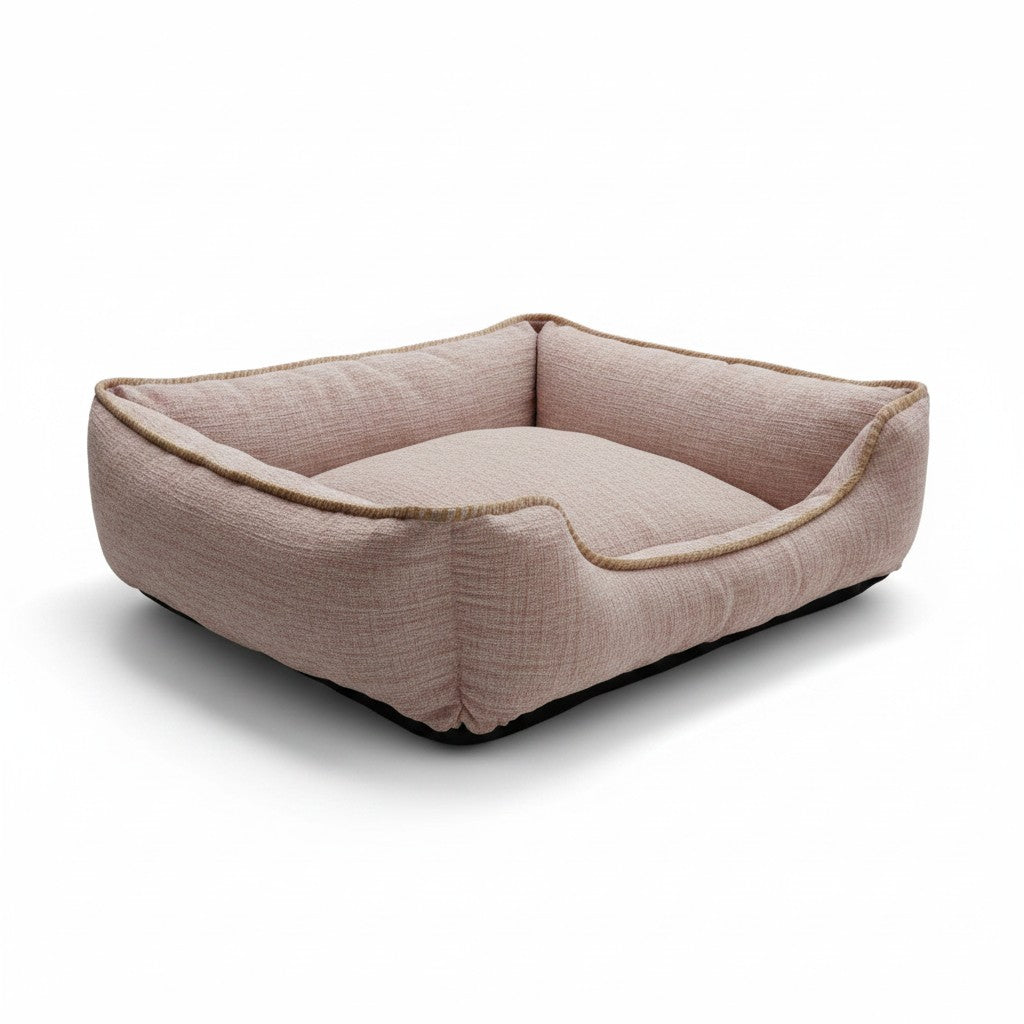 Un panier pour chien de forme rectangulaire, photographié de trois-quarts sur un fond blanc neutre. Le couchage est confectionné dans un tissu texturé de couleur rose poudré chiné, avec des rebords épais et rembourrés pour le confort de l'animal. Le rebord avant est légèrement rabaissé pour faciliter l'accès, et tout le contour supérieur est souligné par une bordure en tissu beige ou marron clair. La base du panier est noire et antidérapante.