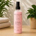Flacon spray rose de 250 ml ANJU Beauté Total Force Lotion Kératine pour chiens et chats, présenté sur une table en bois avec des serviettes beiges et une plante verte en arrière-plan.